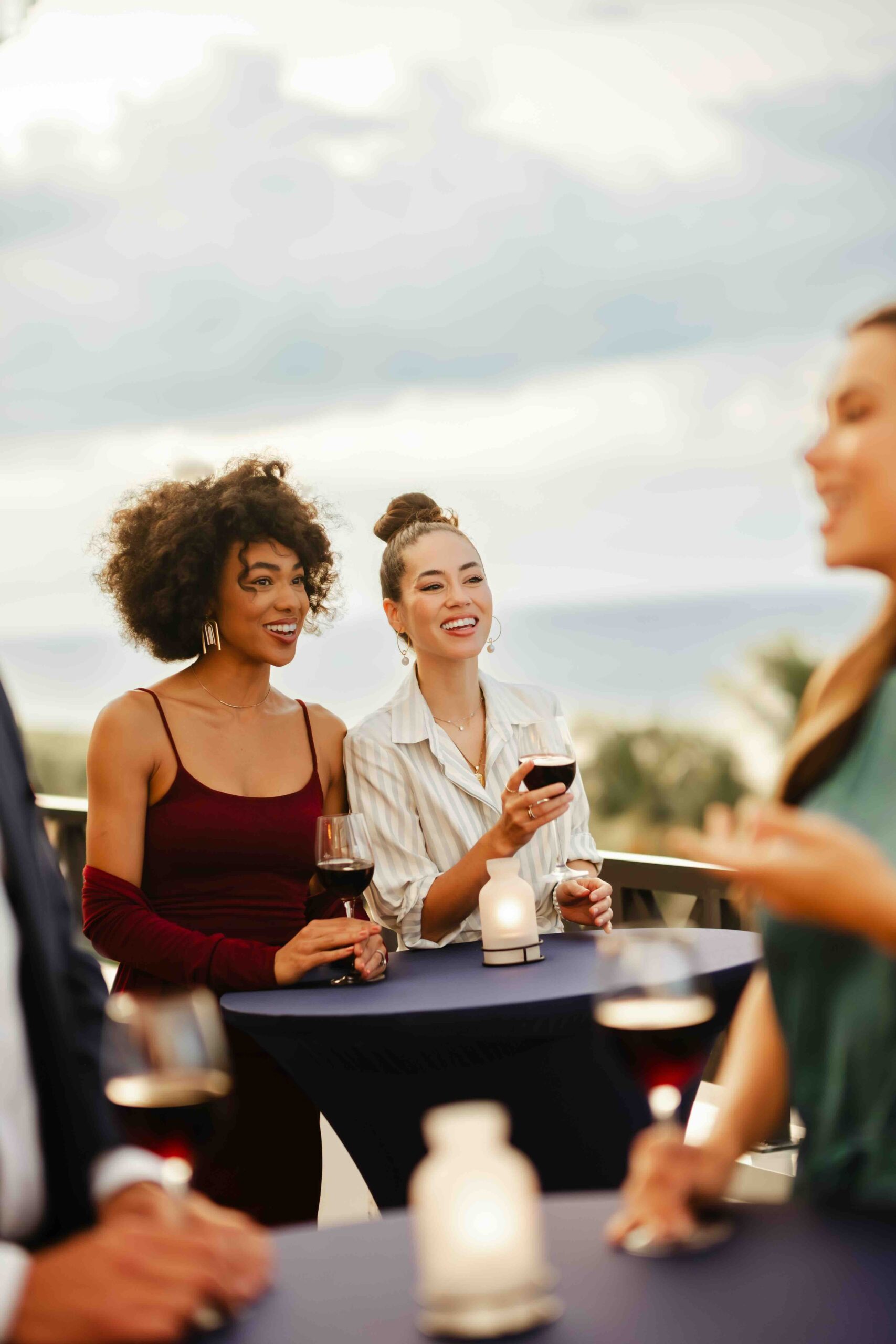 Two women stand at a cocktail table outdoors, smiling and holding glasses of wine. One wears a burgundy dress, the other a white blouse. The setting appears to be a casual social gathering, with candles on the tables.