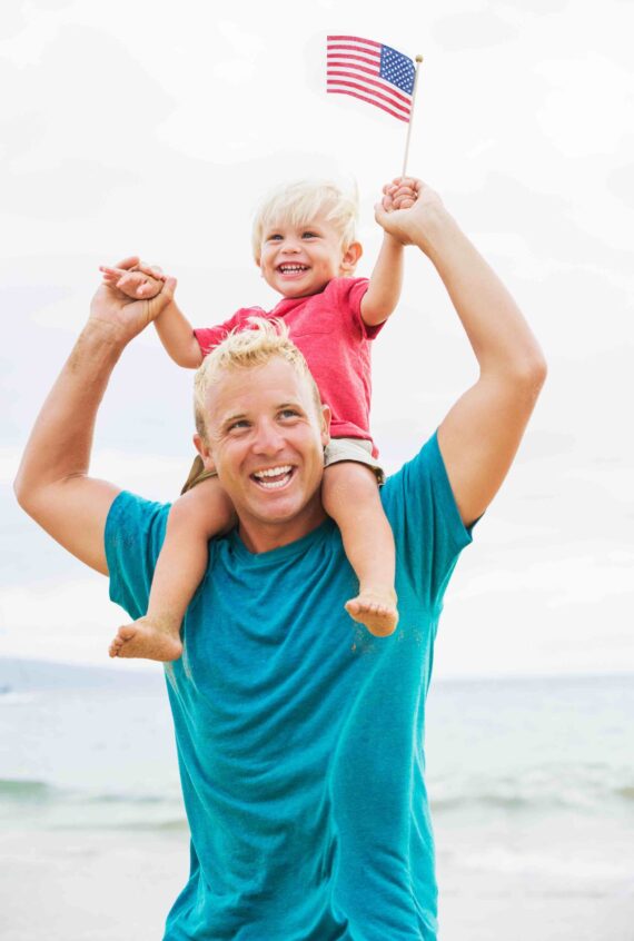 A smiling man gives a young boy a piggyback ride on a beach. The boy, also smiling, holds a small American flag up high. Both have blonde hair and are enjoying a bright, cheerful day outdoors.
