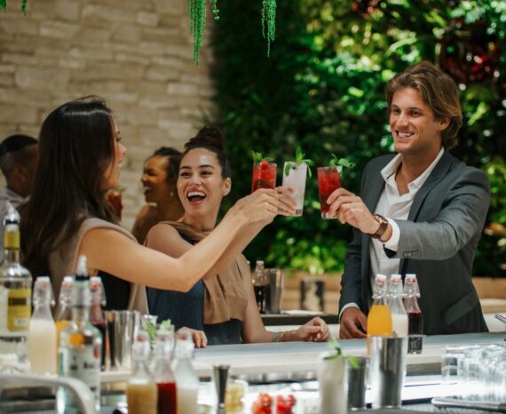 Three people at a modern bar clink colorful cocktails together, smiling and celebrating. Bottles and glasses are on the counter, with a green plant wall in the background.