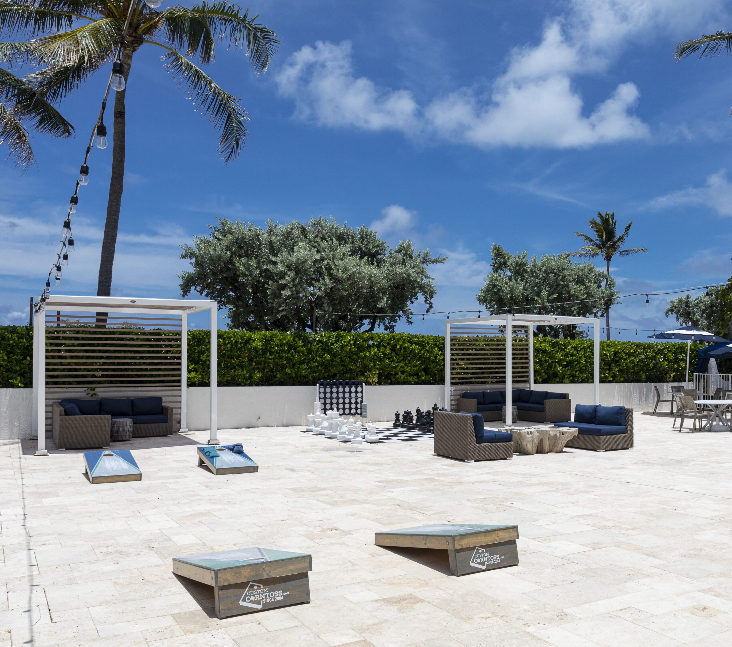 Outdoor lounge area with sofas under pergolas, palm trees, string lights, cornhole boards, and a large chess set on a sunny day with blue sky and clouds.