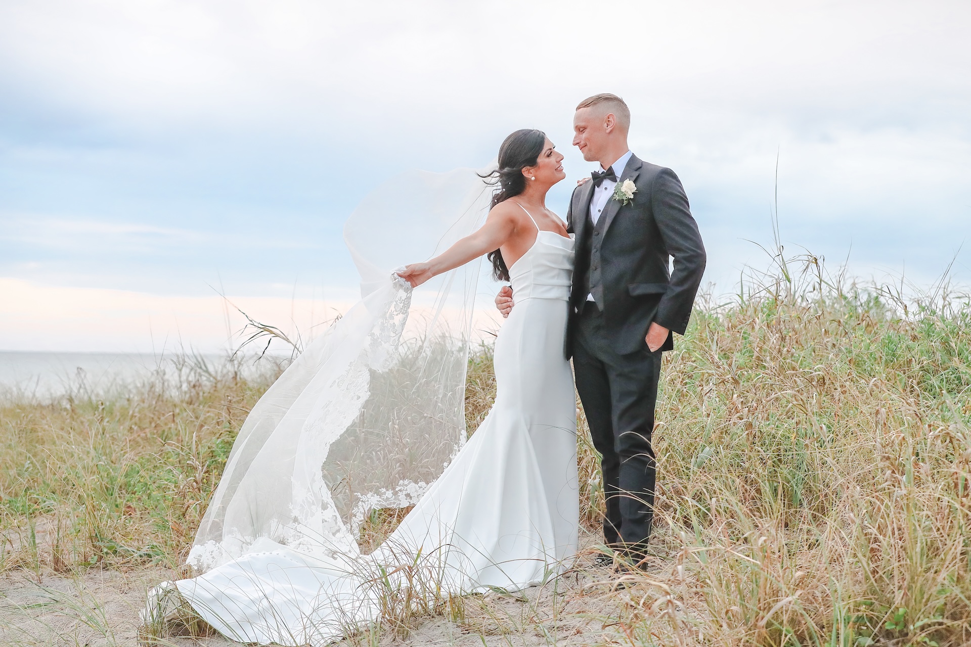 A bride and groom stand on a beach, gazing at each other. The bride holds her veil flowing in the wind, and both are dressed in formal wedding attire, surrounded by tall grass and a cloudy sky.