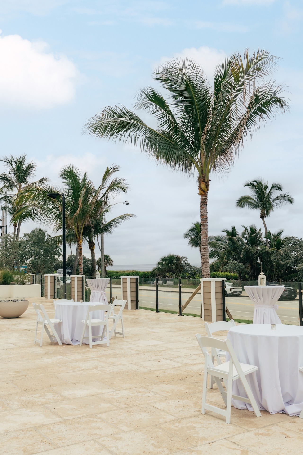An outdoor patio with palm trees, round cocktail tables draped in white cloths, and white folding chairs, set up for an event under a partly cloudy sky.