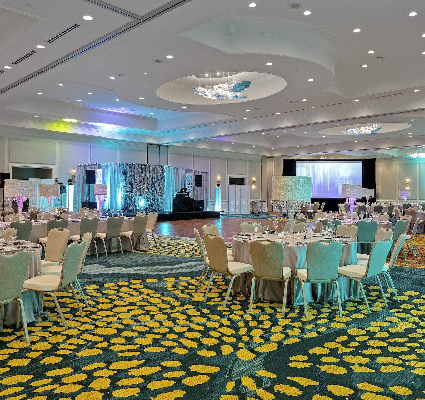 A large, brightly lit banquet hall is set up for an event with round tables, chairs, elegant table settings, and modern lighting in blue and purple tones. A stage and projector screens are visible in the background.