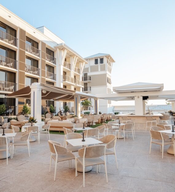 Outdoor terrace at a modern, light-colored hotel with white tables and chairs, shaded seating areas, and a clear blue sky in the background. The ocean is visible beyond the patio.