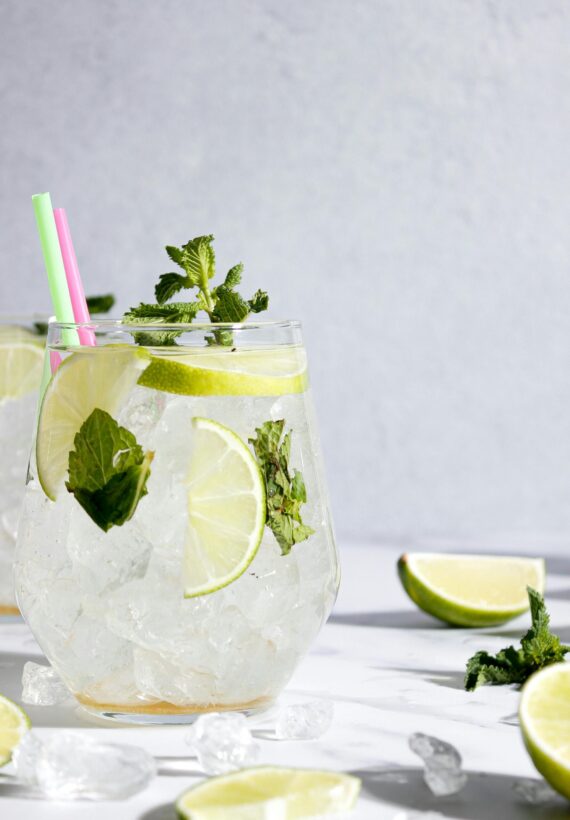 A refreshing drink in a glass filled with ice, garnished with lime slices and mint leaves. Two colorful straws are in the glass. Additional lime wedges and mint are scattered around on a light surface.