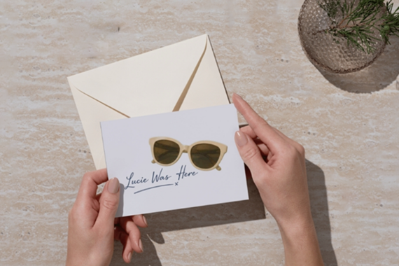 A person holds a card showing sunglasses and the handwritten words Lucie Was Here x, with an envelope on a beige stone surface and a glass dish with a small plant in the corner.