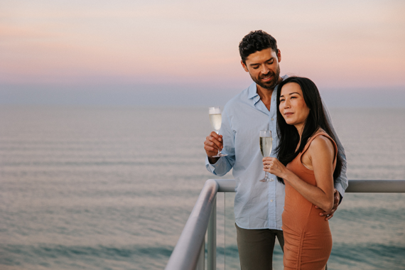 A couple stands on a balcony by the ocean at sunset, holding champagne glasses and smiling, with calm water and a pastel sky in the background.