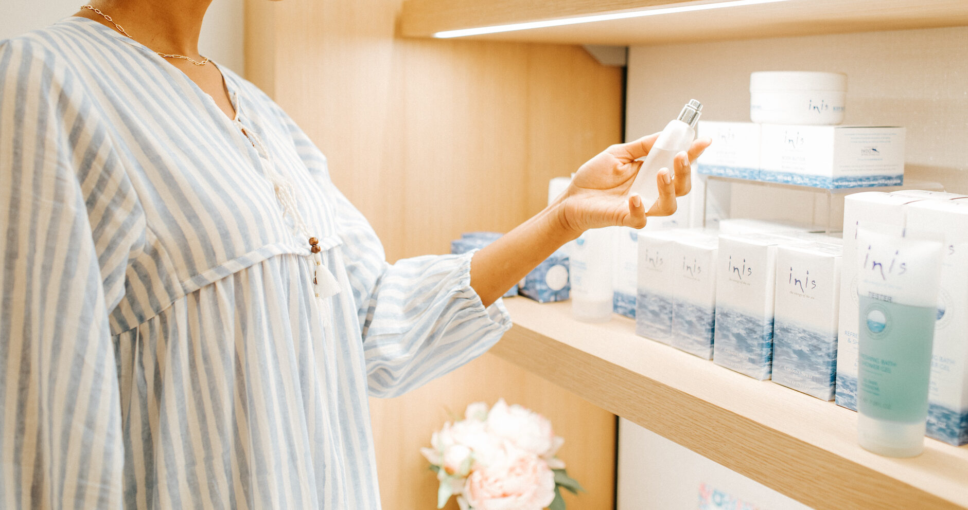 A person in a striped blouse examines a small bottle from a display shelf filled with skincare products. There are boxes and jars labeled Inis beside a vase with pink flowers in the background.