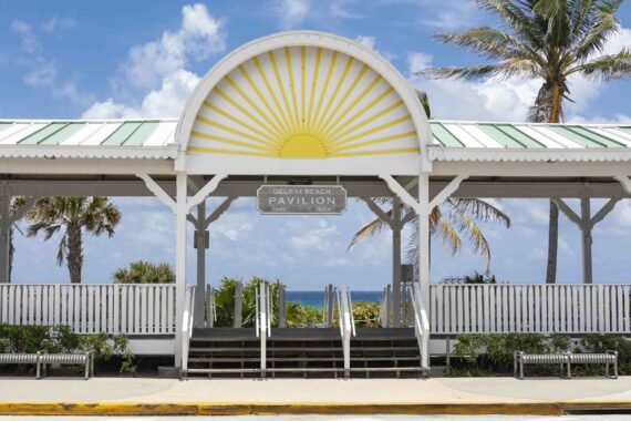 A white pavilion at Delray Beach with a sunburst design on the arch. It is framed by palm trees and benches, with the ocean visible in the distance. The sky is partly cloudy. A sign reads Delray Beach Pavilion.