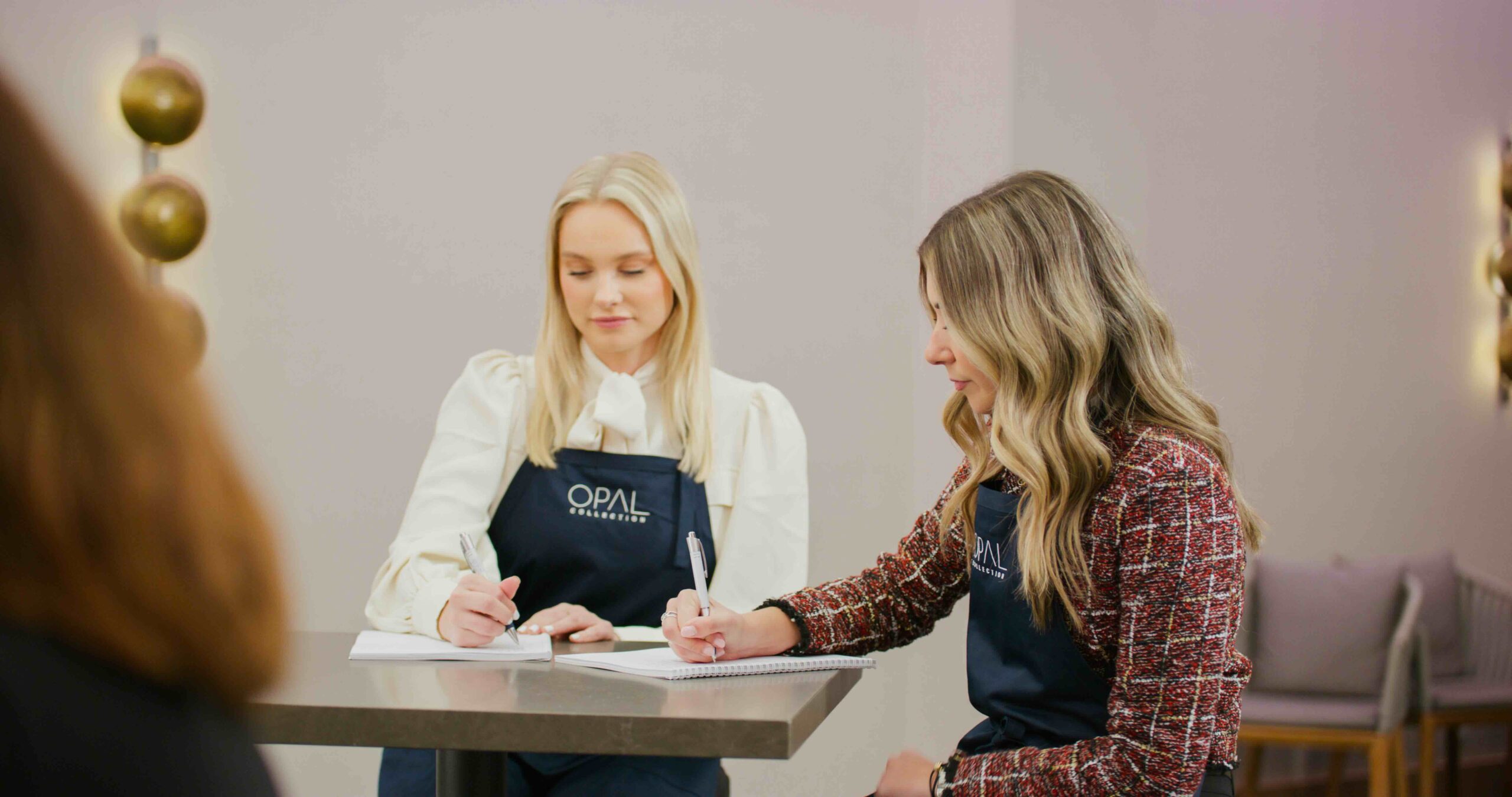Two women wearing dark aprons sit at a table writing in notebooks. They appear to be in a bright, modern indoor setting, focused on their work.