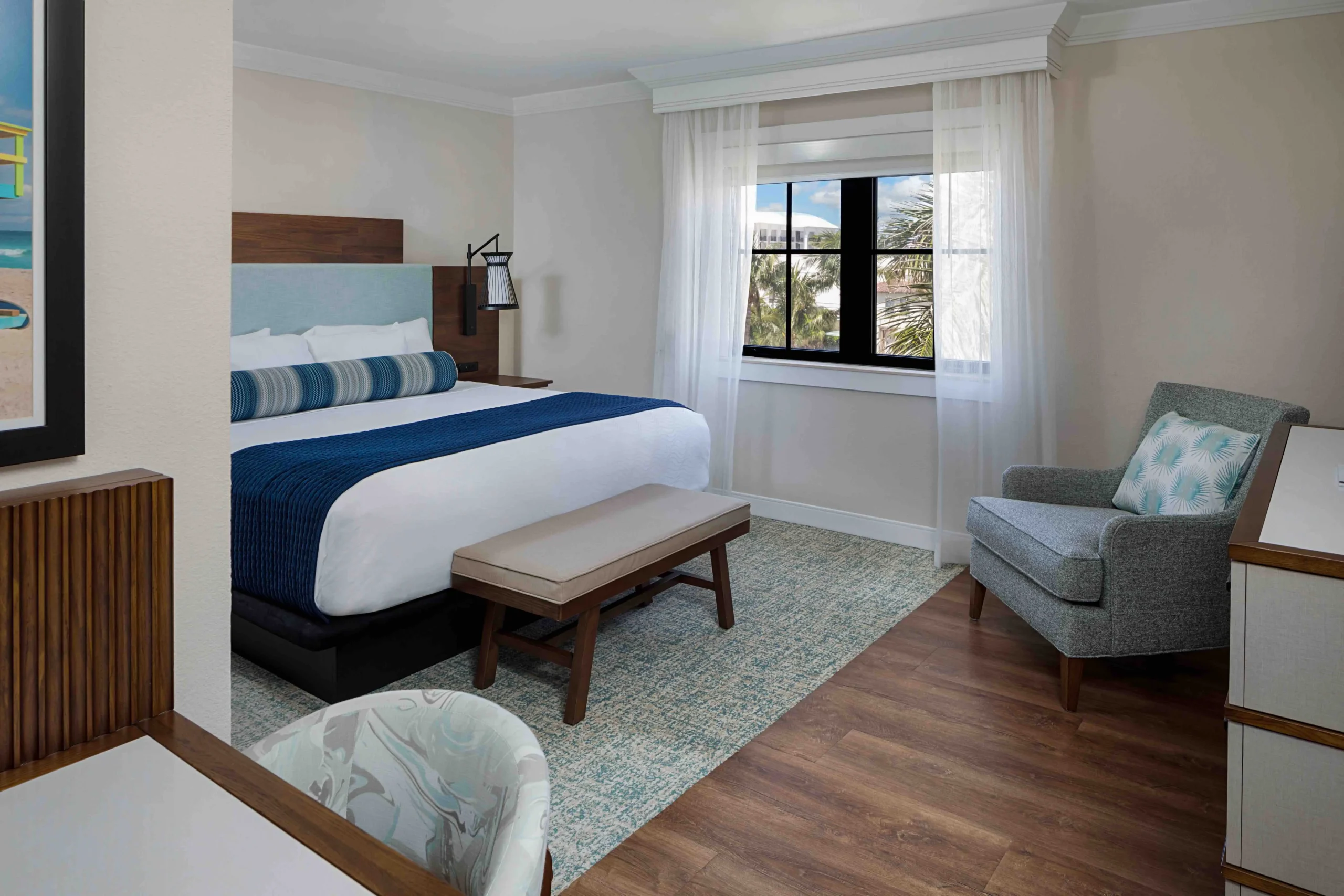 A modern hotel room features a neatly made bed with blue and white bedding, a bench at the foot, an upholstered chair by the window, wood floors, and light walls. A framed picture and desk are also visible.
