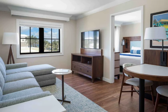 A modern hotel suite with a gray sectional sofa, wall-mounted TV, wooden furniture, a small dining table with chairs, and a bedroom visible through an open door, featuring a bed with blue and white bedding.