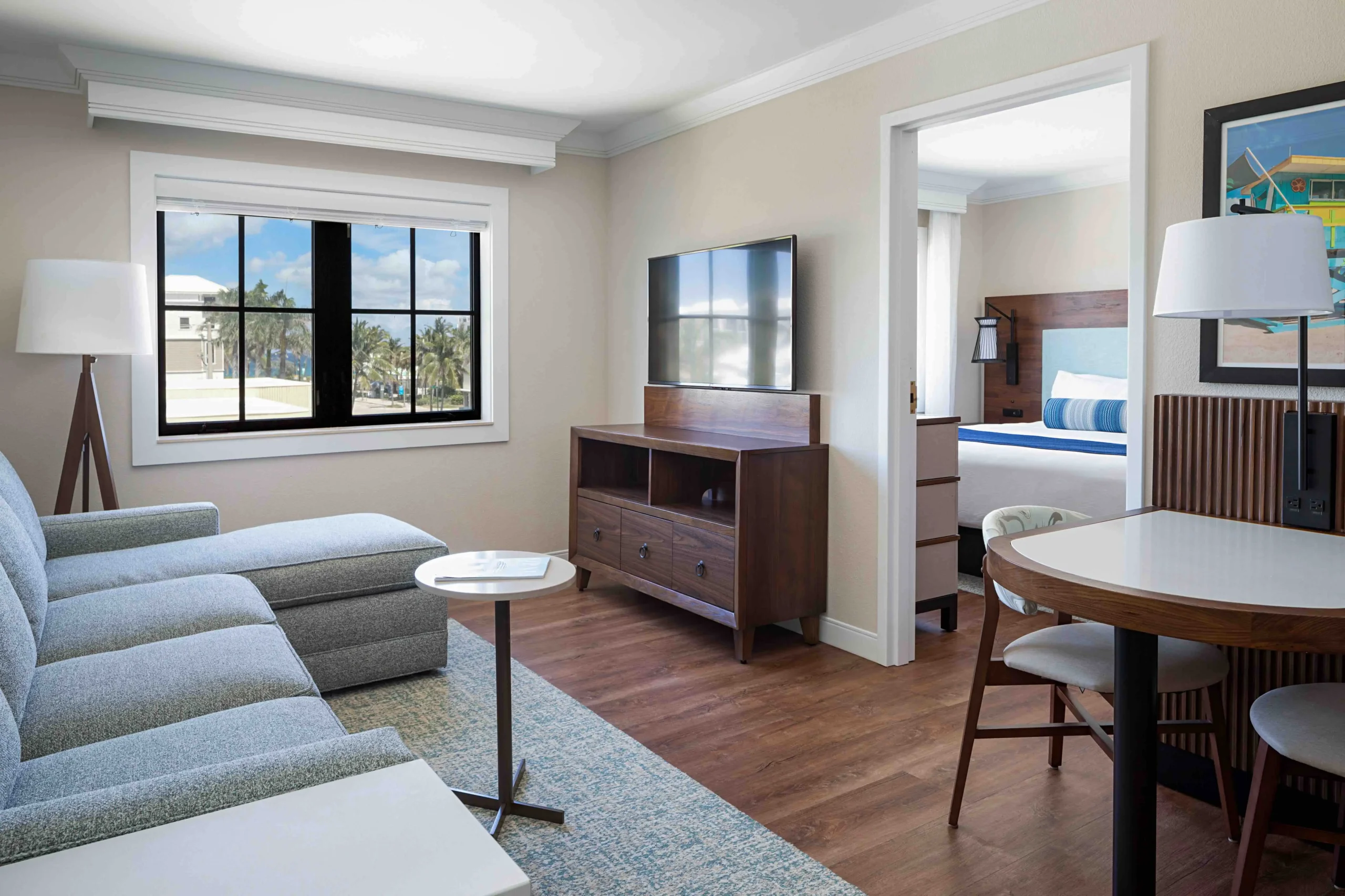 A modern hotel suite with a gray sectional sofa, wall-mounted TV, wooden furniture, a small dining table with chairs, and a bedroom visible through an open door, featuring a bed with blue and white bedding.