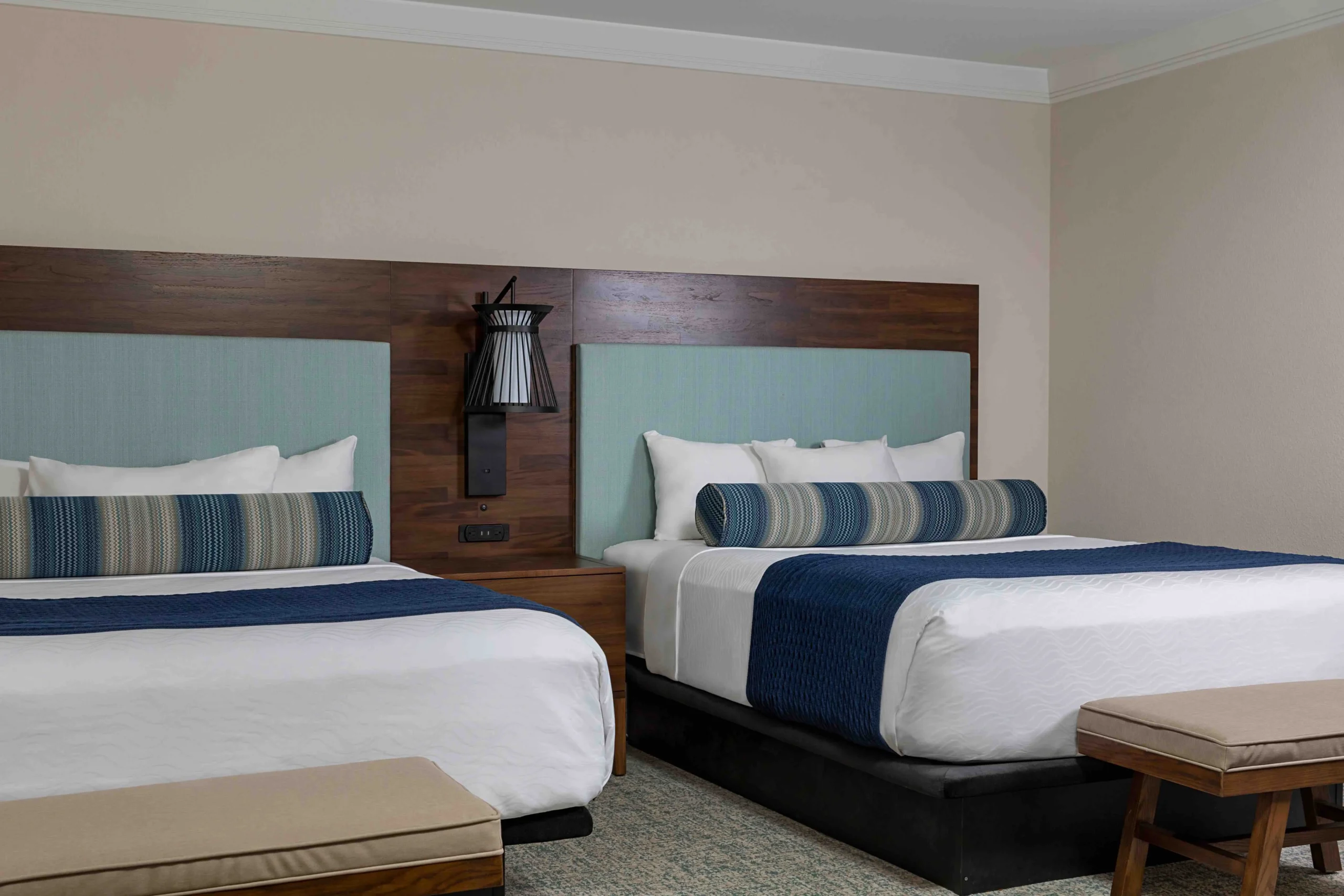 Hotel room with two double beds, each with white linens, blue accent blankets, and striped bolster pillows; wooden headboards, a wall lamp, and a wooden bench and chair are also visible.