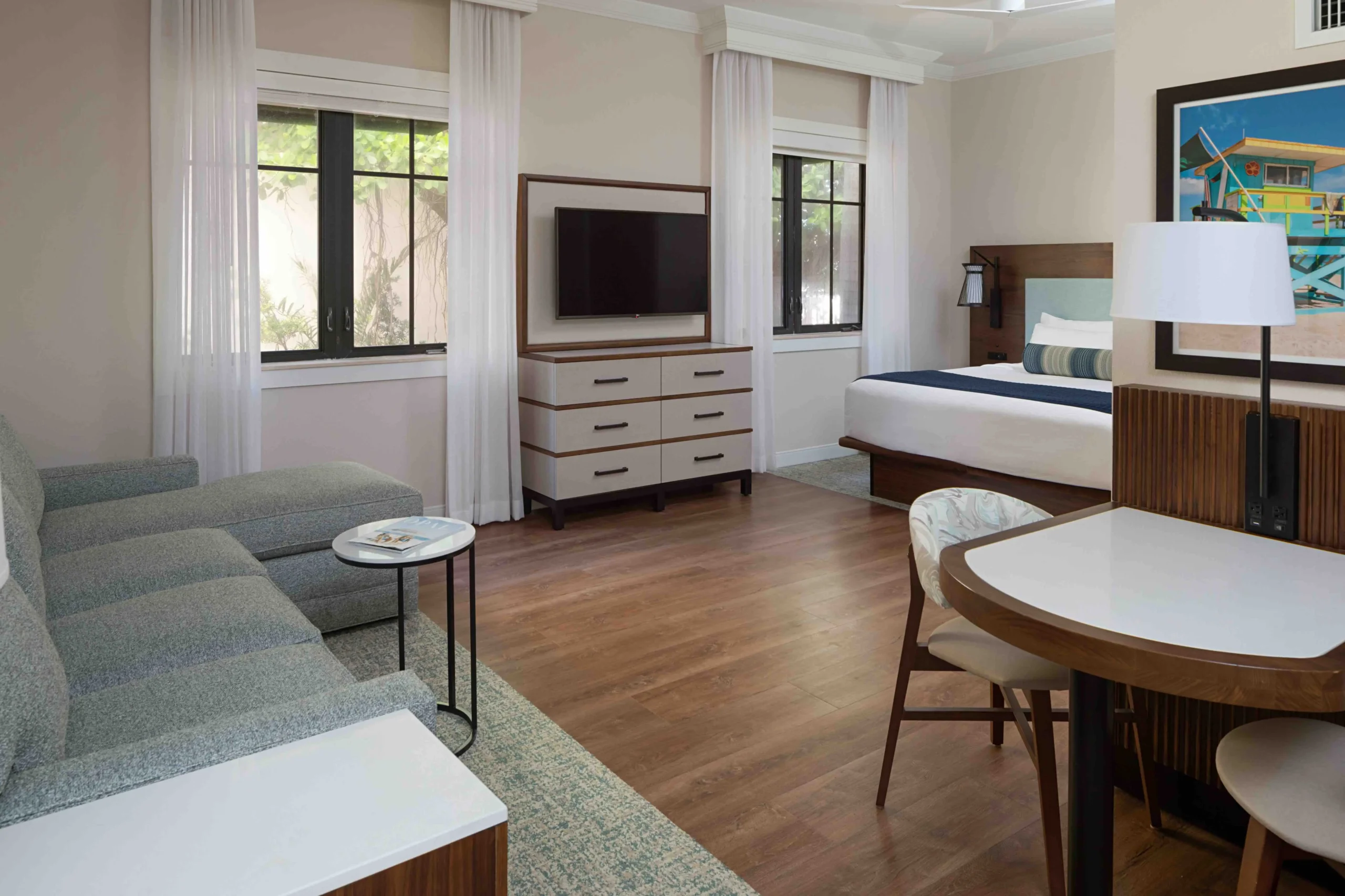 A modern hotel room with wood flooring, a bed, a gray sectional sofa, a flat-screen TV on a dresser, a round dining table with chairs, and large windows with white curtains.