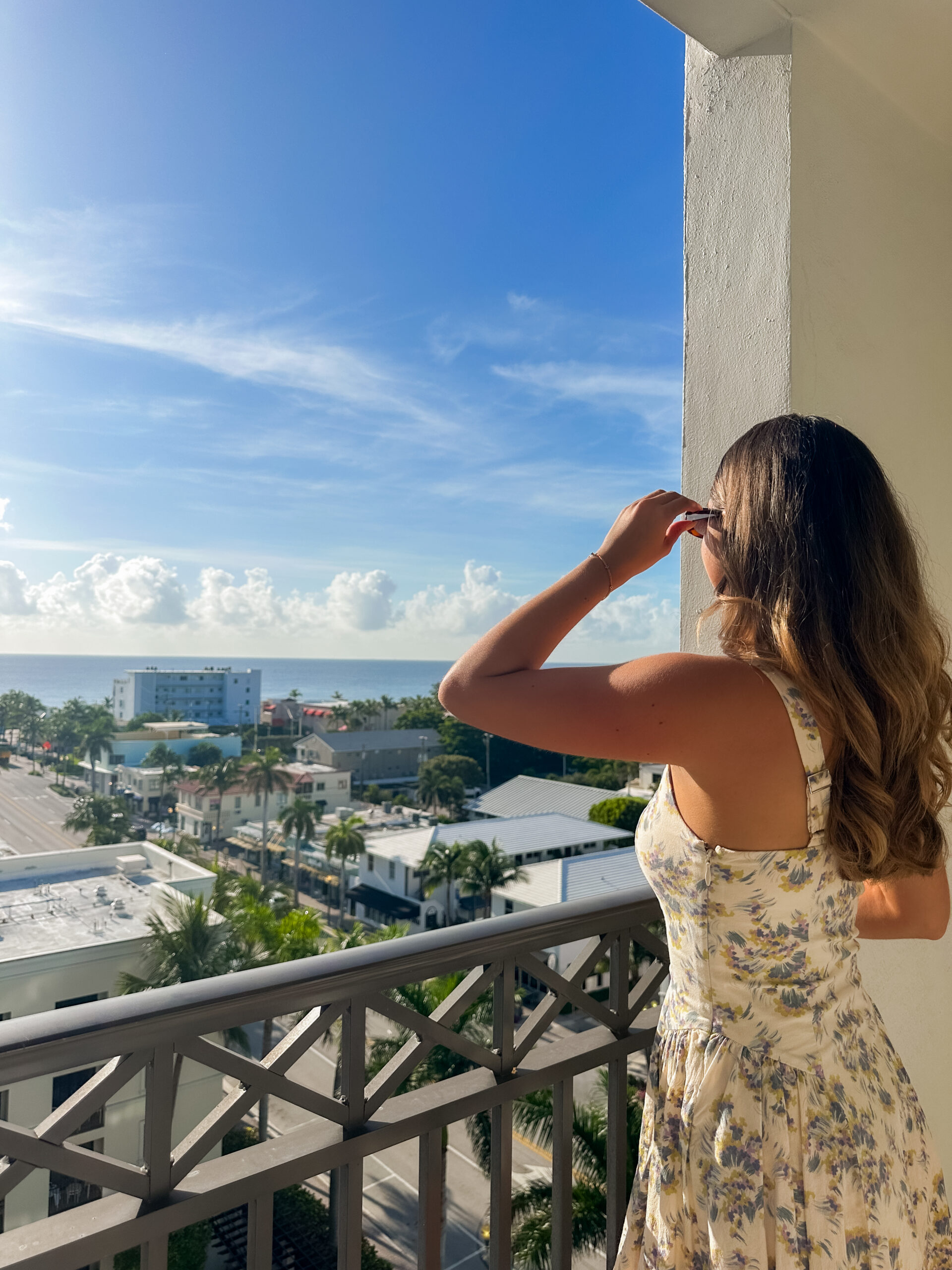A woman in a floral dress stands on a balcony, looking out at a sunny coastal cityscape with palm trees, buildings, and a blue sky filled with clouds.