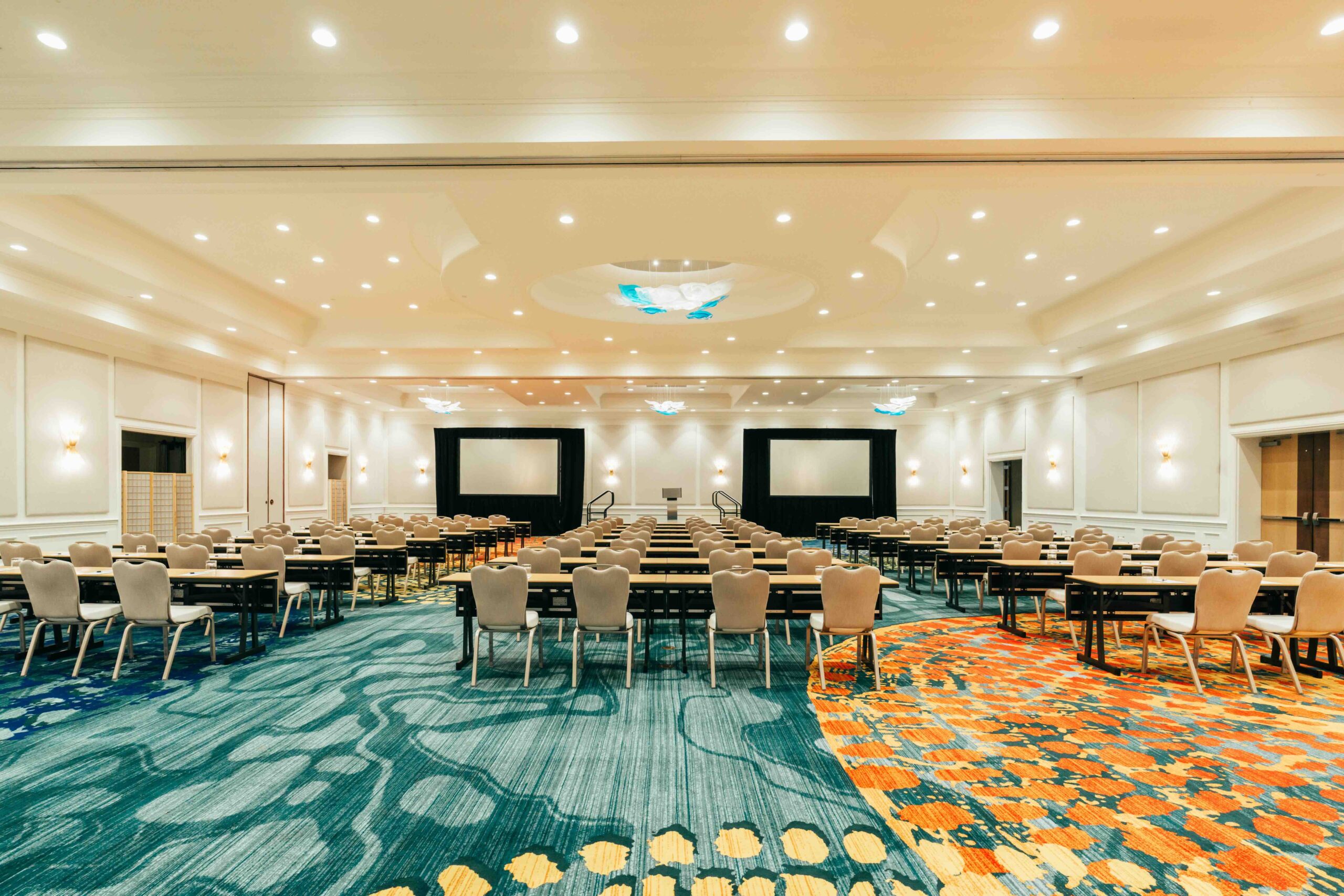 A spacious, well-lit conference room with rows of tables and chairs facing three large projection screens. The room features colorful patterned carpet and modern ceiling lights.