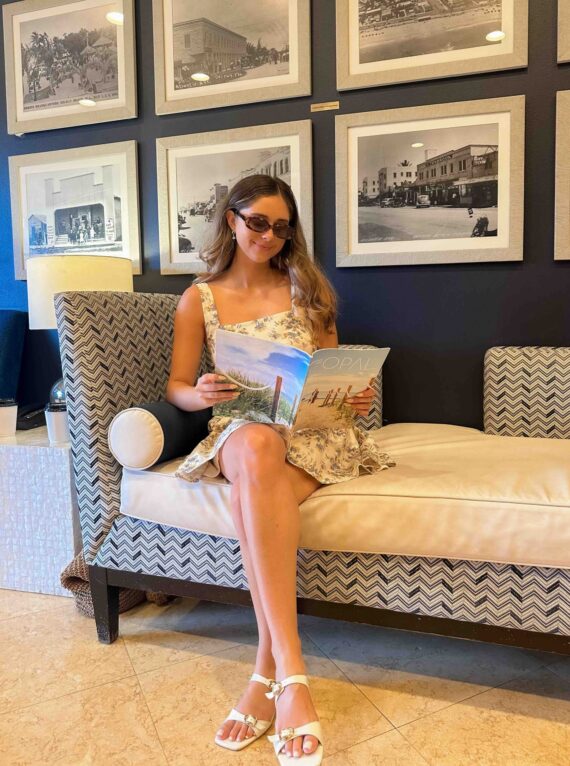 A woman in a patterned summer dress and sunglasses sits on a cushioned sofa reading a magazine, with black-and-white photos displayed on the wall behind her.