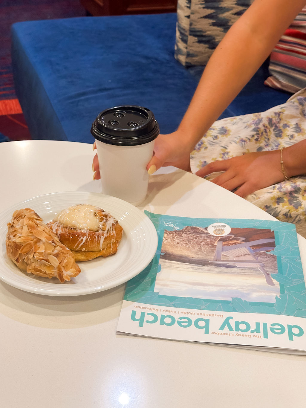 A person reaches for a white takeaway coffee cup on a table with a pastry and an almond-covered treat on a plate, next to a turquoise magazine titled Delray Beach.