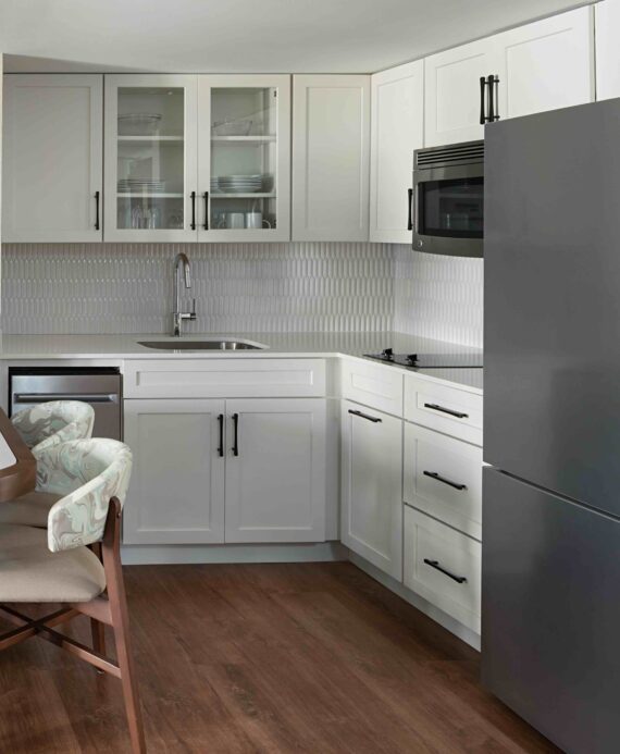 Modern kitchen with white cabinets, stainless steel sink, microwave, gray refrigerator, and patterned tile backsplash. Wooden floor and dining chair are visible in the foreground.