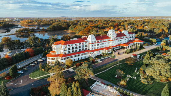 Aerial view of a large, elegant hotel with red roofs surrounded by trees with autumn foliage. Theres a winding driveway with cars parked and a well-maintained lawn. A scenic body of water and hills can be seen in the background under a blue sky.