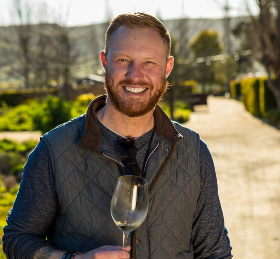 A smiling man holding a glass of white wine outdoors.