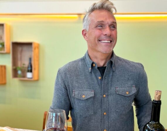 A smiling man holding a glass of red wine at a bar with a bottle of Castello di Rampolla Sangiovese nearby.