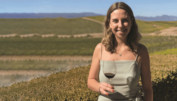 A woman in a light green dress stands outdoors holding a glass of red wine, smiling at the camera with green fields and distant hills in the background under a clear blue sky.