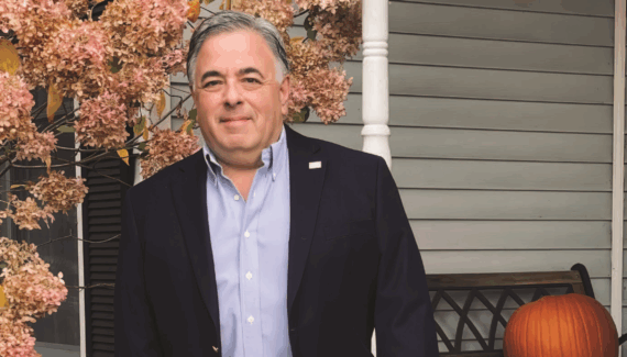 A man in a navy blazer and light blue shirt stands on a porch next to blooming hydrangeas and a pumpkin, with a gray house and a white column in the background.