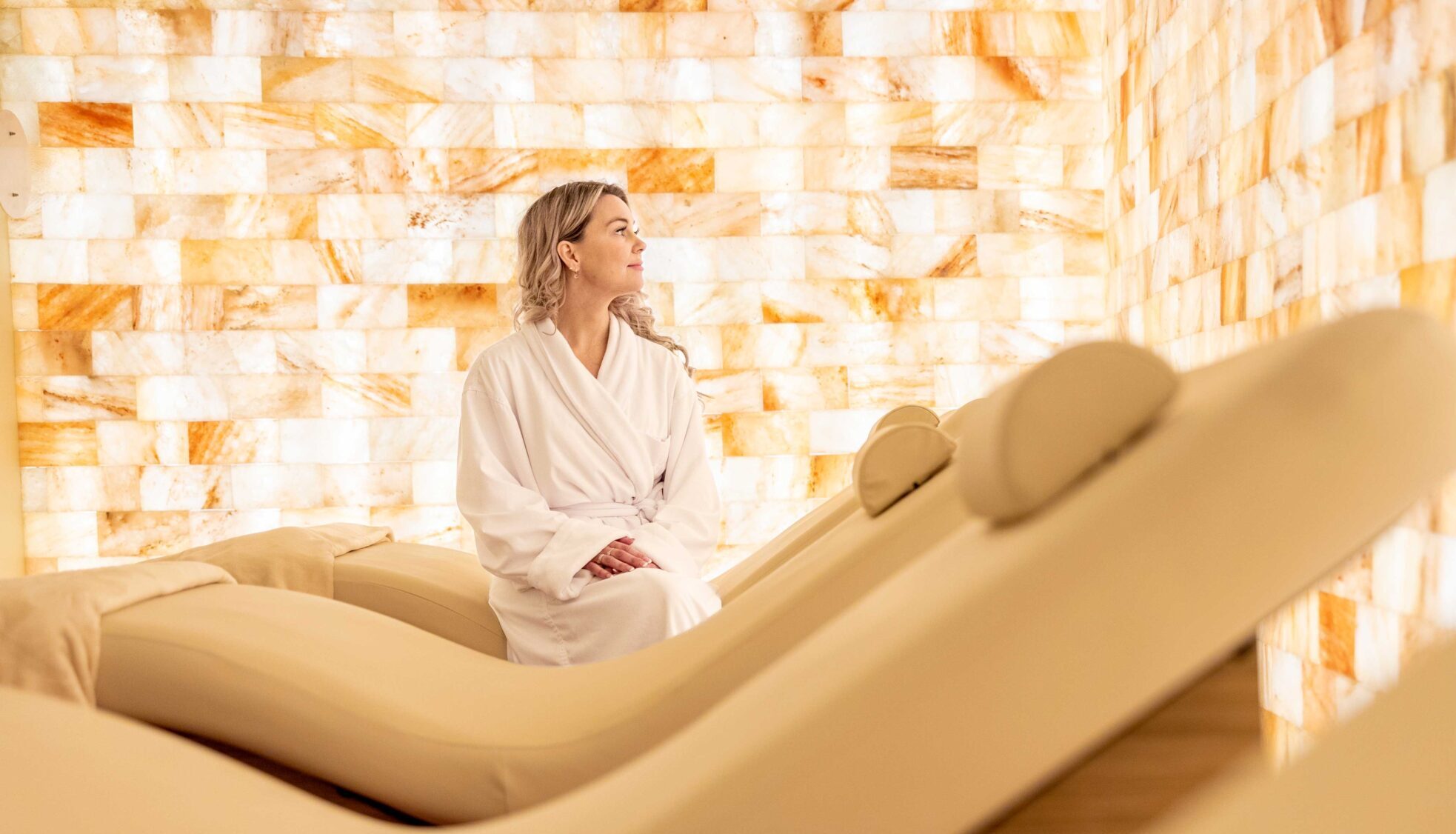 A woman in a white robe sits on a lounge chair in a serene room with warm, glowing walls made of salt bricks. She appears relaxed and contemplative.