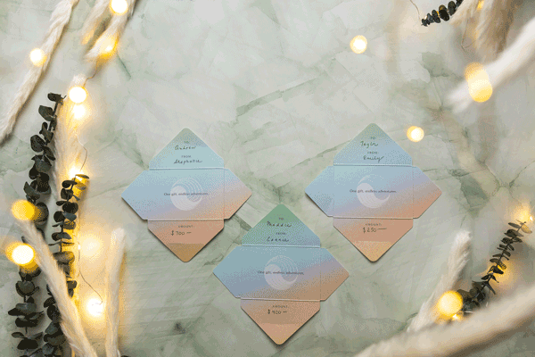 Three pastel-colored envelopes with handwritten notes and gift card amounts are arranged on a marble surface, surrounded by string lights and greenery, creating a cozy, festive atmosphere.