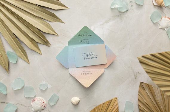 A pastel-colored gift card holder with OPAL COLLECTION branding sits on a marble surface, surrounded by dried palm leaves, sea glass pieces, and small seashells. The card is addressed to Matthew from Audrey for $500.