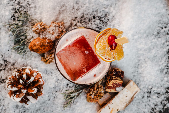 Top-down view of a cocktail with a large square ice cube, garnished with a dried orange slice and a cranberry, surrounded by pine cones and snow.