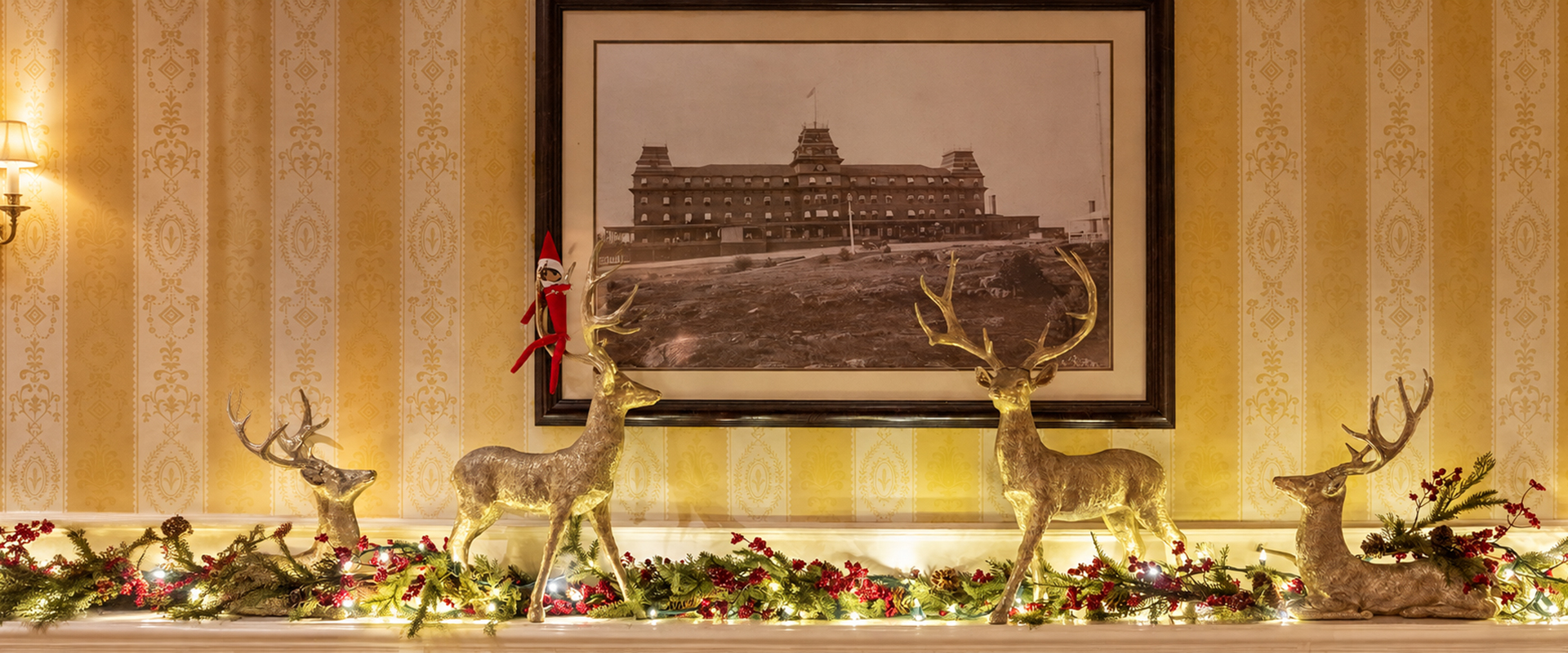 A mantelpiece decorated with four gold deer figurines, Christmas greenery, and a mischievous elf doll, positioned below a framed historical photograph of a large hotel.