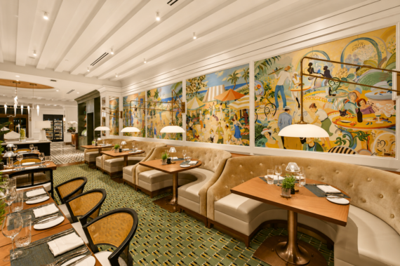 Annie's is an Elegant restaurant interior with plush beige booths, wooden tables set for dining, and a colorful, lively mural spanning the wall, depicting festive outdoor scenes. The room is warmly lit with modern lamps and has a patterned carpet.
