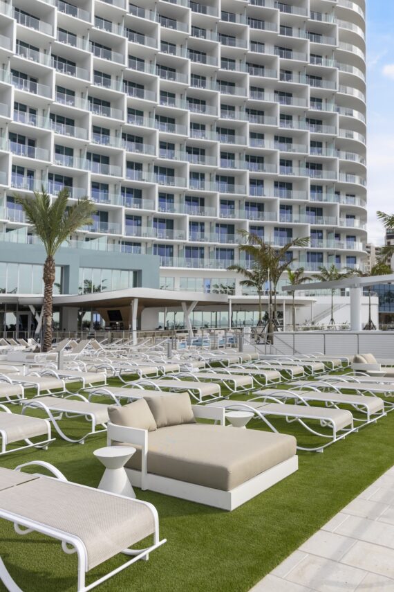 Rows of white lounge chairs and a beige daybed sit on green artificial grass by a modern, curved hotel with balconies. Palm trees are planted near the pool area under a blue sky.