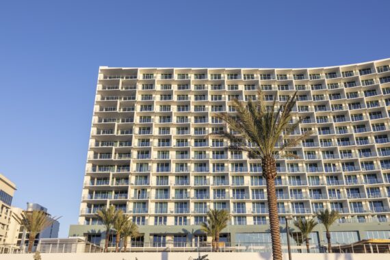 A modern, multi-story hotel building with many balconies and large windows stands under a clear blue sky, with several tall palm trees in front.
