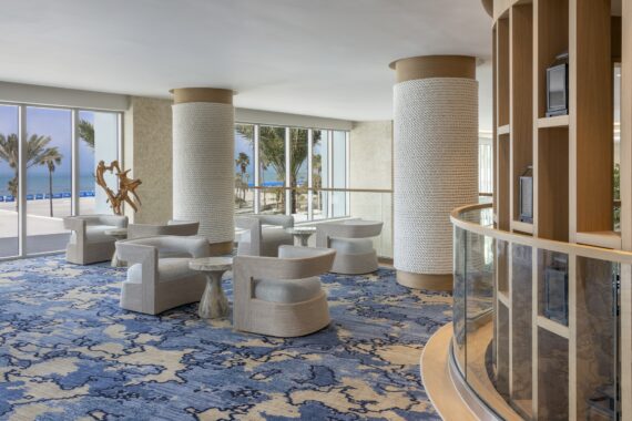 A modern lounge with light-colored chairs and small tables, large windows showcasing palm trees and a beach with blue ocean, and textured pillars on a blue abstract-patterned carpet.