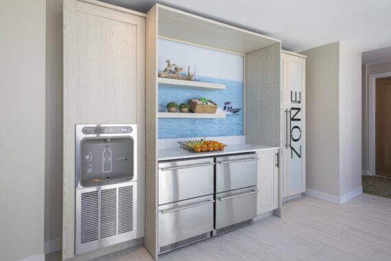 Modern kitchenette with a water dispenser, stainless steel drawers, bowls of oranges, and shelves displaying decorative items against a beach-themed backdrop. Tall cabinets frame the space, with one labeled ZONE.