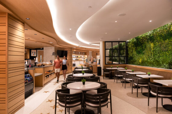 Modern café with wooden accents, round tables, and black chairs. Two people walk past the counter. A lush green vertical garden decorates one wall. The space is bright with curved ceiling lights.