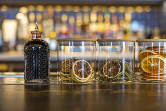 Three glass jars with dried citrus slices and a decorative bottle of dark liquid are arranged on a bar counter with a blurred background of bottles and blue tiles.