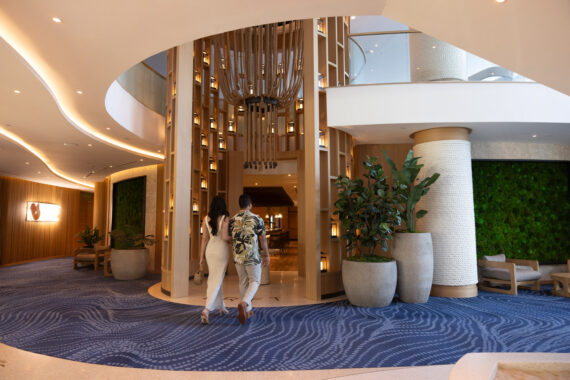 A man and woman walk into a modern hotel lobby with blue patterned carpet, large potted plants, wood accents, and a green living wall. Warm lighting and curved architectural elements create a welcoming atmosphere.