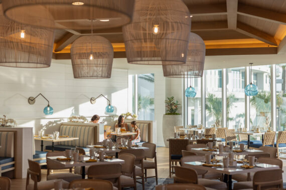 A bright, modern restaurant with large wicker light fixtures, wooden ceiling beams, and floor-to-ceiling windows. Two women are seated at a booth, while other tables and chairs are neatly set but empty.