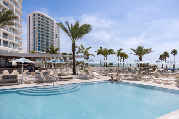 A modern beachfront resort with a large outdoor pool, palm trees, lounge chairs, and umbrellas. Two people relax at the pools edge, with a high-rise hotel and ocean visible in the background under a blue sky.