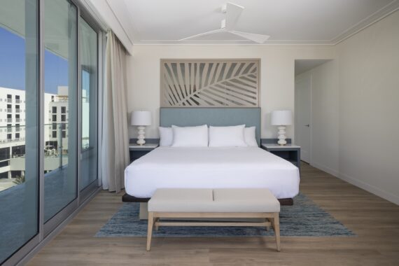 A modern hotel room with a large bed, blue headboard, two bedside tables with lamps, a patterned rug, a small bench at the foot of the bed, and floor-to-ceiling windows with city views.