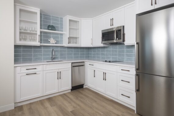 Modern kitchen with white cabinets, stainless steel appliances, light wood flooring, a blue tiled backsplash, and open shelves displaying glassware and decorative items.