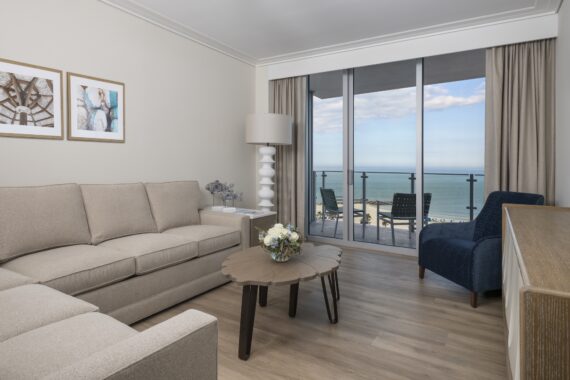 A modern living room with beige sofas, a blue armchair, a wooden coffee table, and large windows showing a balcony with ocean views under a partly cloudy sky. Framed art and neutral decor complete the space.