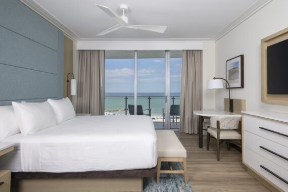 Modern hotel room with a large bed, blue accent wall, desk, TV, ceiling fan, and floor-to-ceiling windows showing a balcony and ocean view under a partly cloudy sky.