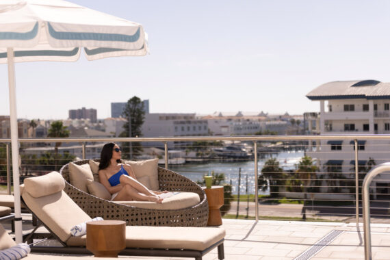 A woman in a blue swimsuit relaxes on a round outdoor lounge chair under an umbrella, overlooking a marina at Clearwater Beach, enjoying the sunshine—perfect for those taking advantage of a Florida resident hotel deal.