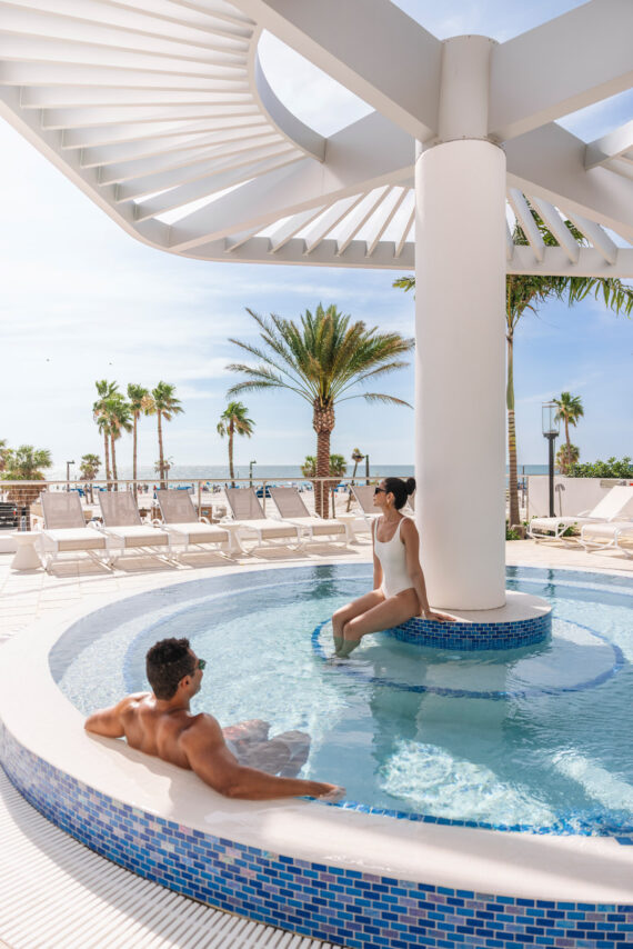 A man and a woman relax in a small circular pool under a modern white shade structure, surrounded by palm trees and empty lounge chairs on a sunny day.